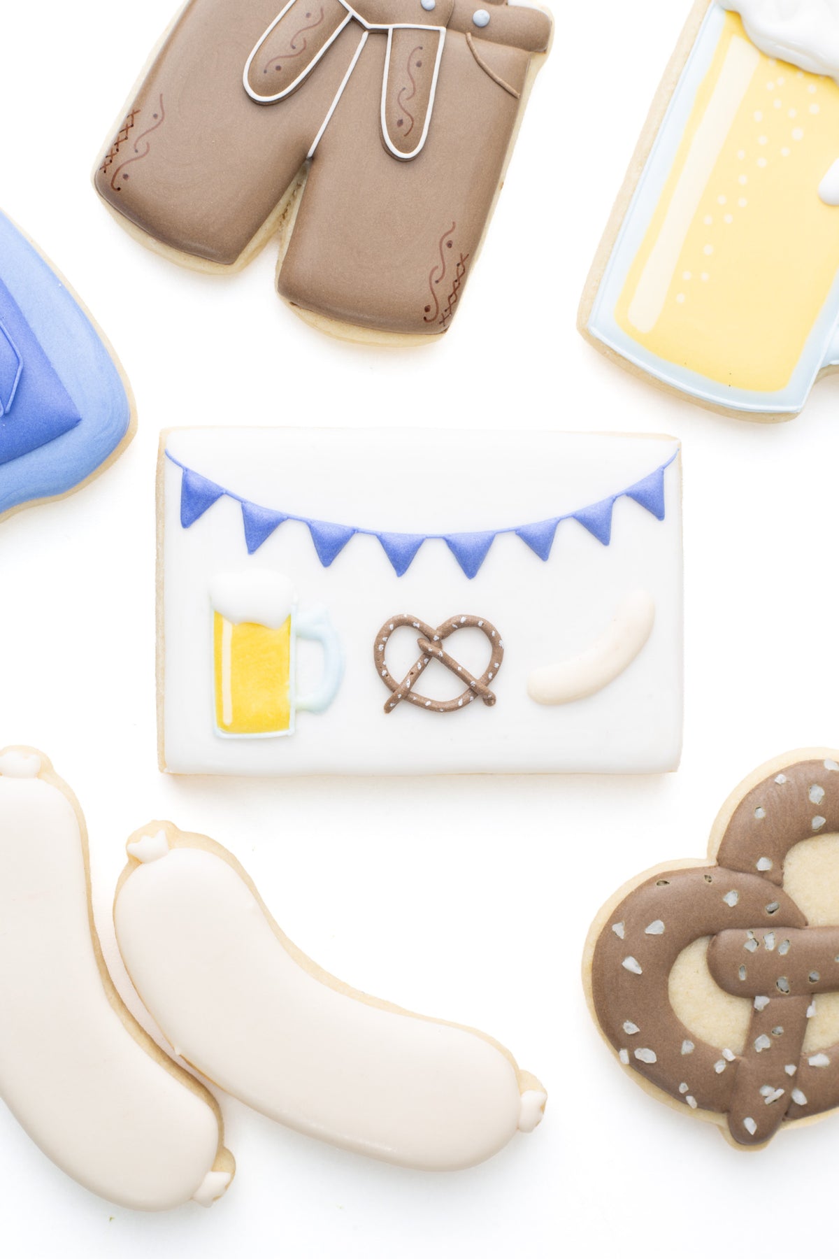 Oktoberfest themed cookies. A square plaque decorated sugar cookie with a beer mug, pretzel, and bratwurst next to a lederhosen, beer mug, bratwurst, and pretzel decorated sugar cookies.