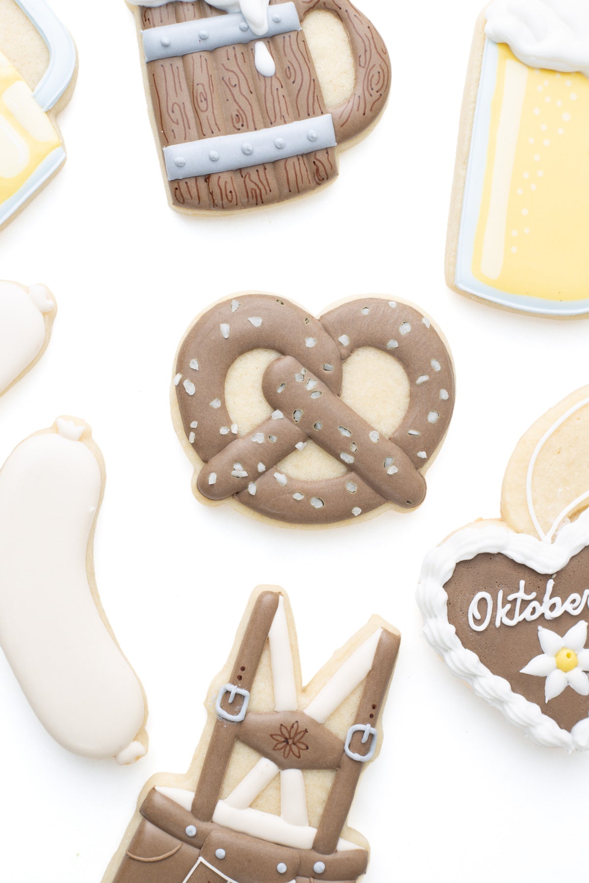 Oktoberfest themed cookies. A pretzel decorated sugar cookie next to a wood beer stein, beer mug, Lebkuchen, lederhosen, bratwurst and dimple beer mug decorated sugar cookie.