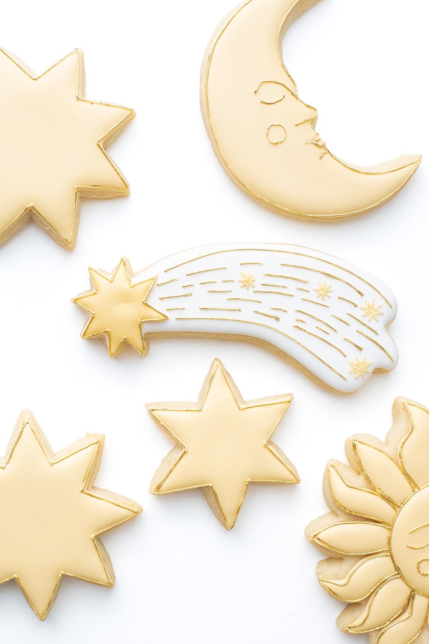 A shooting star decorated sugar cookie next to a celestial star and crescent moon with face and a 6 point,7 point, and 8 point star.