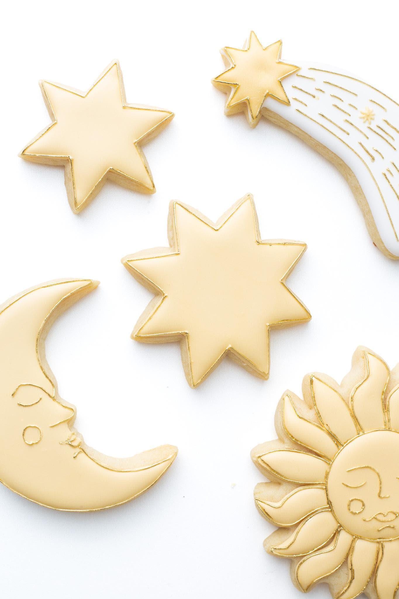 A 6 and 8 point star  decorated sugar cookie next to a celestial crescent moon and sun with a face and shooting star.