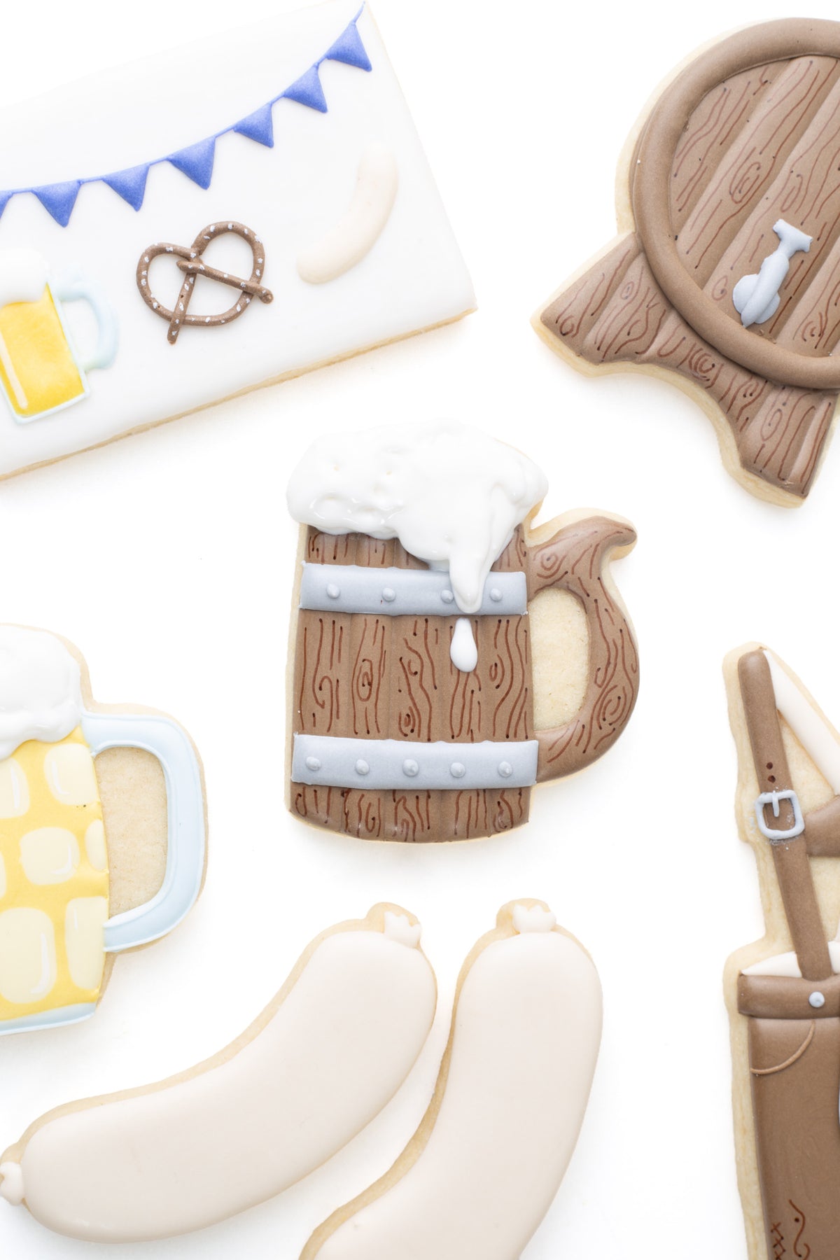 Oktoberfest themed cookies. A wood beer mug decorated sugar cookie next to a ,  beer stein, wood beer stein, keg, pretzel and bratwurst decorated sugar cookie.